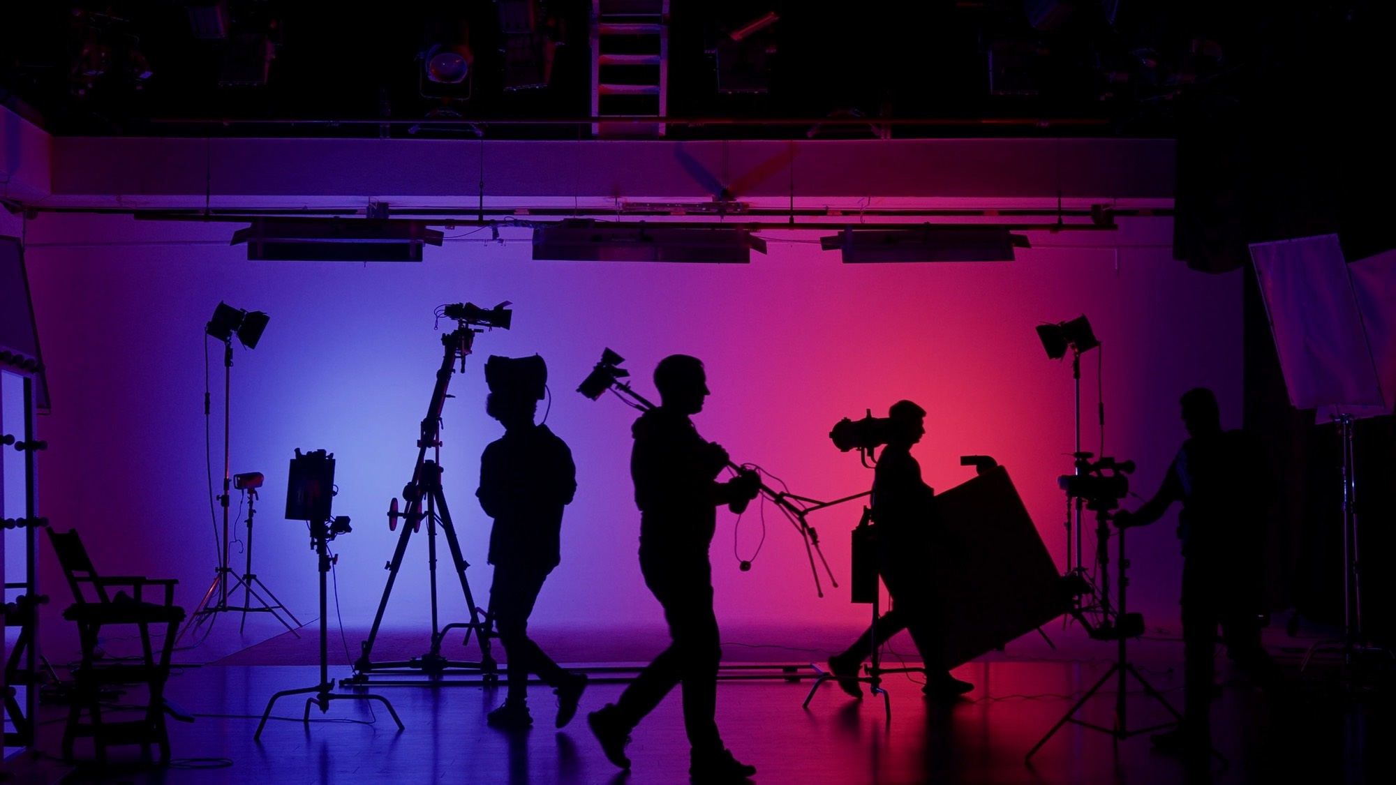 Silhouetted camera crew and production equipment cross a studio lit in blue and magenta