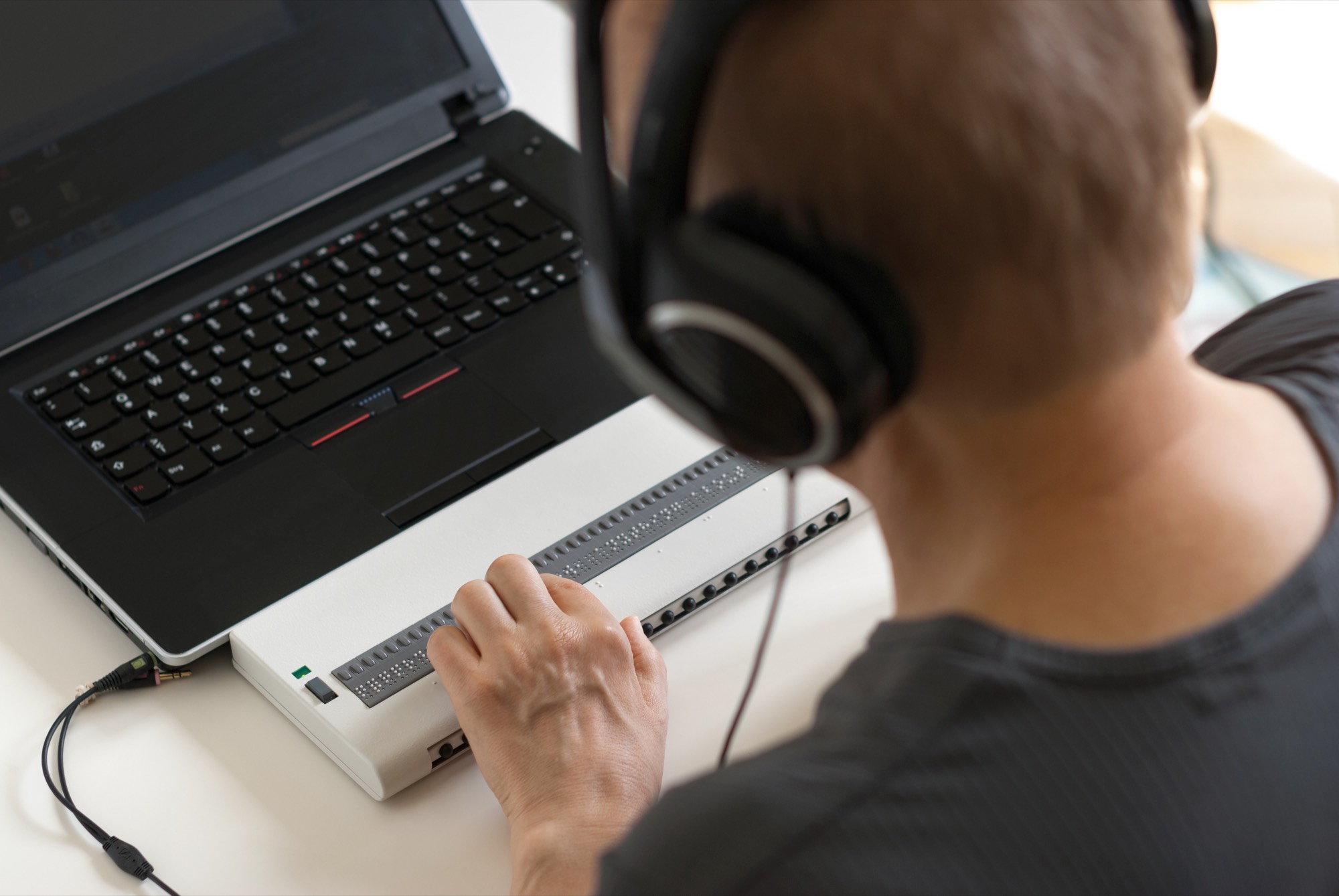 A blind computer user wears headphones and reads a refreshable Braille display beside a laptop