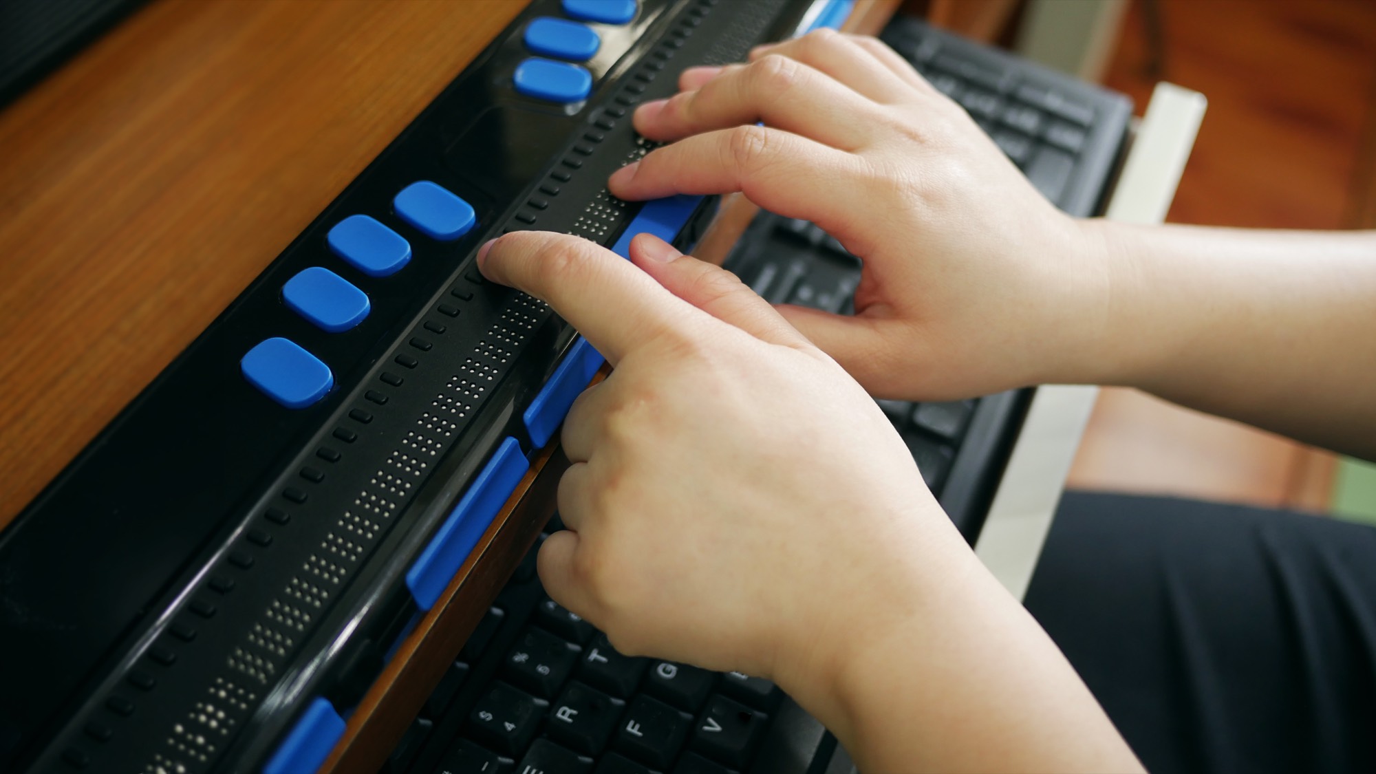 Hands read a refreshable Braille display positioned above a computer keyboard