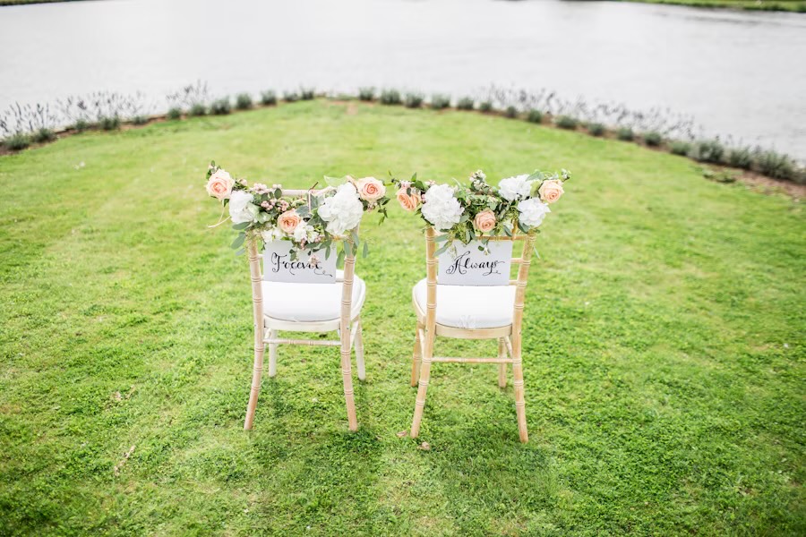 Two flower-trimmed chairs by a lake, their backs lettered Forever and Always.