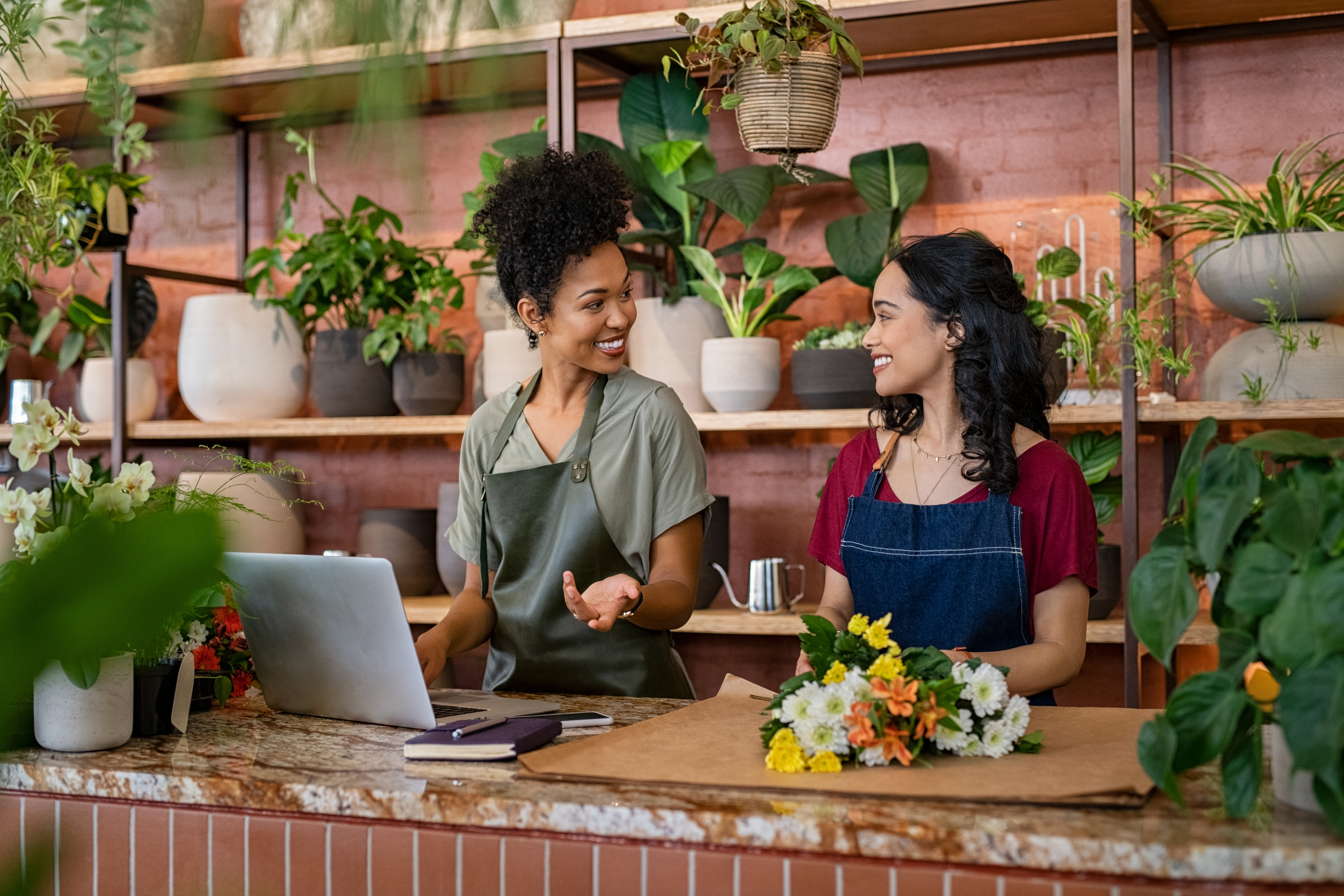 A flower-shop owner works on a laptop behind a counter surrounded by flowers
